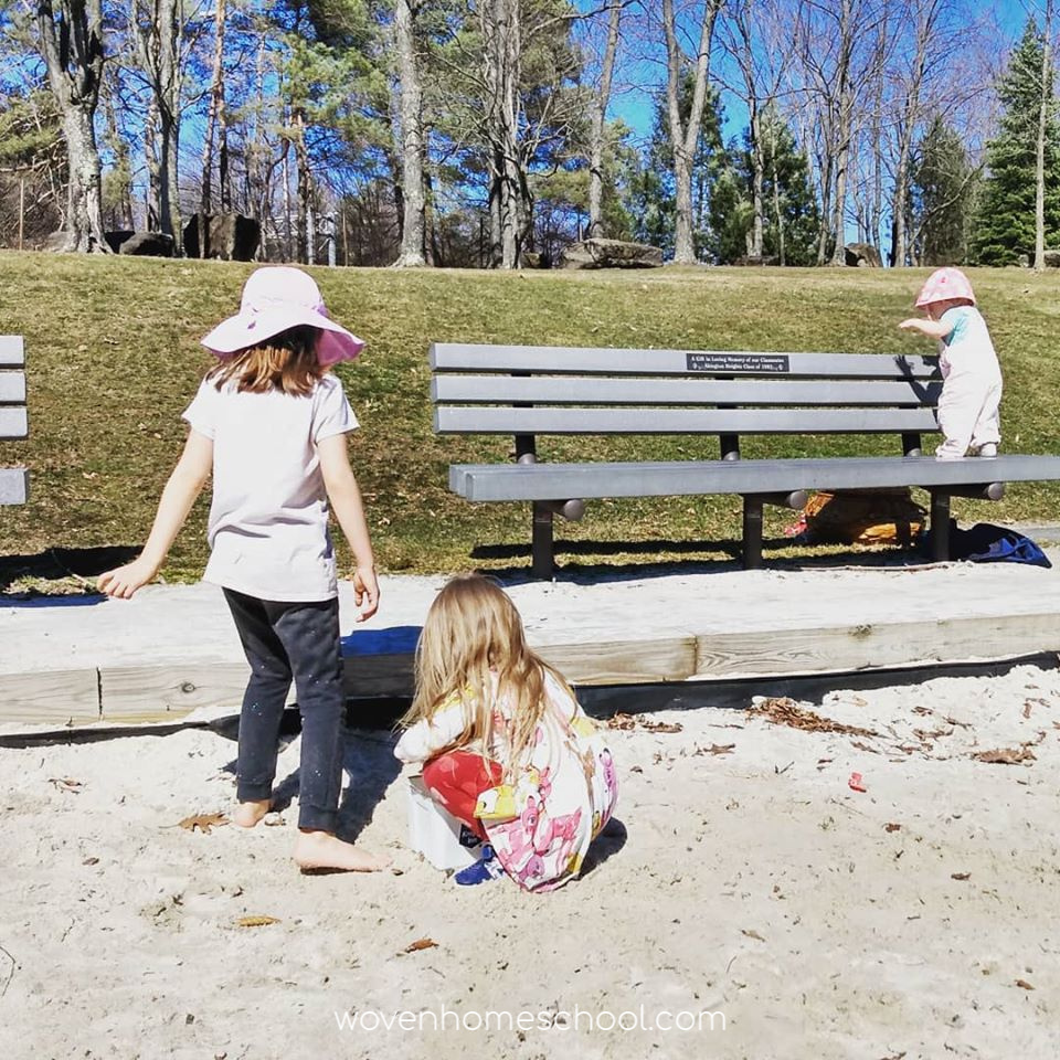 Two girls playing in a sandbox with a toddler standing on a bench nearby.